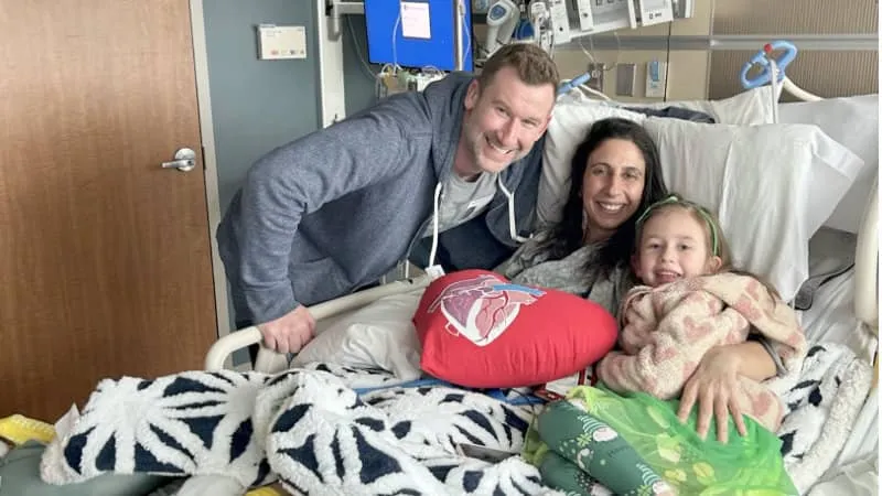 Kristin Fludder laying down on a hospital bed smiling with her husband and daughter by her side.
